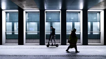 Man rides scooter, other person walks; nighttime urban environment.