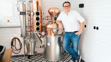 Man stands relaxed next to a copper still in a modern setting.