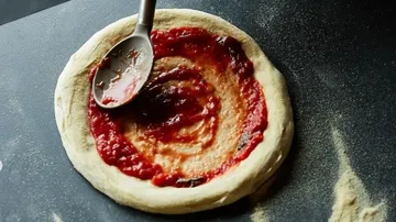 A pizza base is coated with sauce on a work surface.