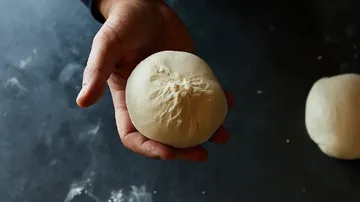 One hand holds a dough ball over a work surface.