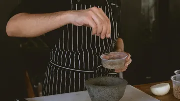 Person in gestreifter Schürze würzt Zutaten in einer Küche, hält Gewürze über einem Mörser, Vorbereitung auf das Kochen.