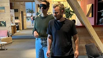 A blindfolded woman walks in a room with a man.
