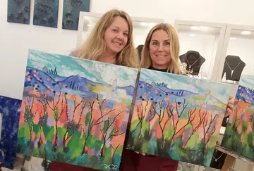 Two women hold colorful paintings in front of an exhibition wall.