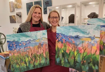 Two women smile next to their colorful paintings in a studio.