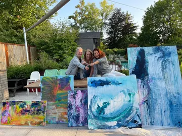 Three women pose in front of colorful paintings in the garden.