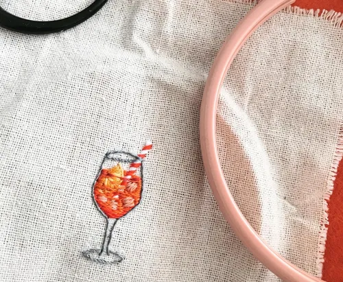 Ein gesticktes Glas mit orangefarbenem Getränk auf Stoff.