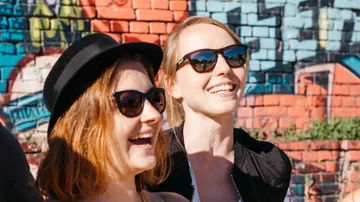 Two women laugh in front of a colorful graffiti wall.