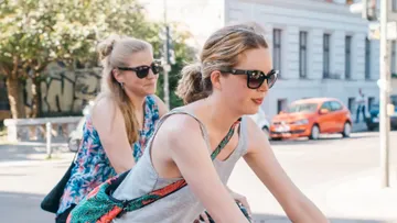 Two women ride bikes in an urban environment.
