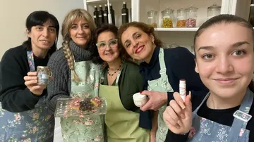 Five women smiling, holding food items in kitchen.