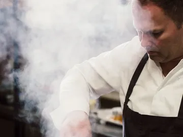 Cook stirs in steam surrounded by a kitchen environment.
