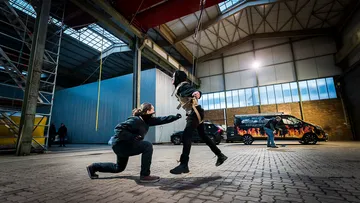 Person suspendiert, eine weitere kniend, Lagerhalle.