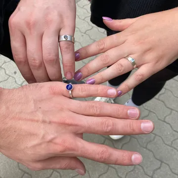 Zwei Hände zeigen Ringe, mit farbigen Nägeln, auf Asphalt.