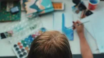 Junge malt mit Pinsel auf Papier, umgeben von Malutensilien.