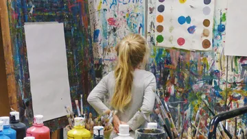 A girl stands in front of a colorful painting wall and thinks.