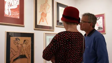 Zwei Personen betrachten Kunstwerke mit Aktzeichnungen in einem Ausstellungsraum, die Kunstwerke sind in verschiedenen Rahmen an der Wand arrangiert.