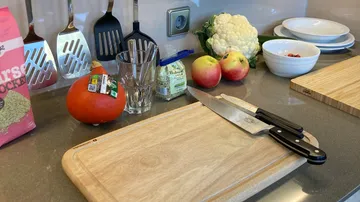 Ein Holzbrett mit Messern, Gemüse und Äpfeln auf einer Theke.