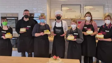 Six people present cakes in a kitchen.