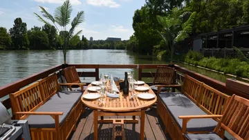 Holztisch mit Stühlen, gedeckt, am Fluss im Freien.