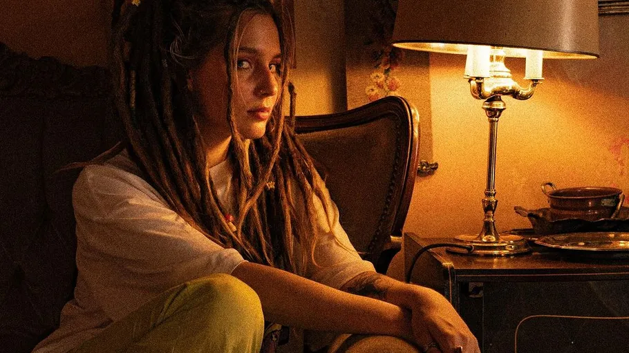 Boy with dreadlocks sits next to a lamp in a room.