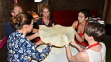 Six people stretch out a large dough together.