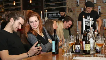 Young people sit at a table, look at cell phones, drink beer.