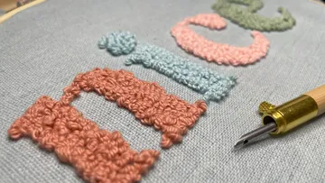 Embroidered letters lie on fabric, tools next to it.