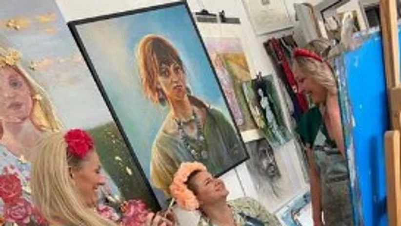 Three women laugh and pose in a studio with works of art.