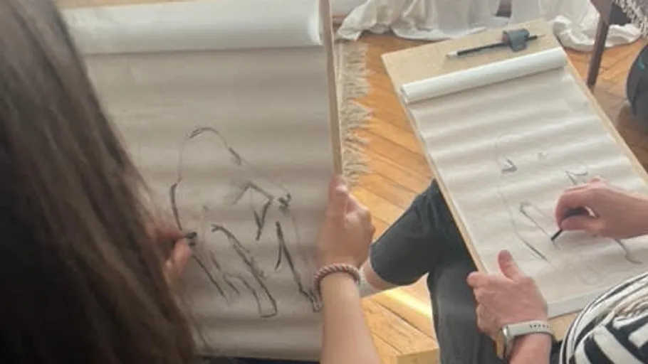 Two people draw on paper surrounded by wooden floors.