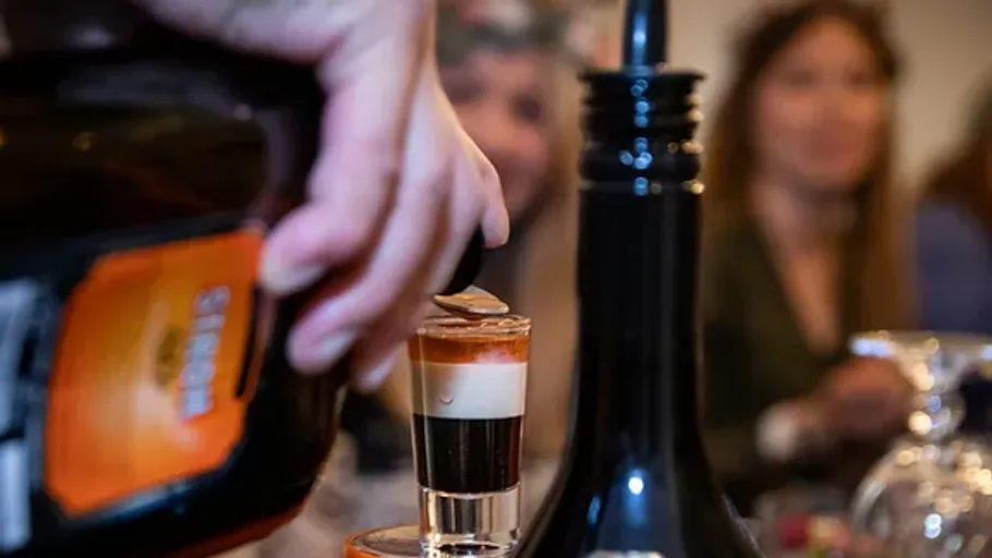 A glass of layered drink is poured, people laugh.