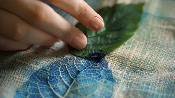 A hand touches a leaf on a blue fabric surface.