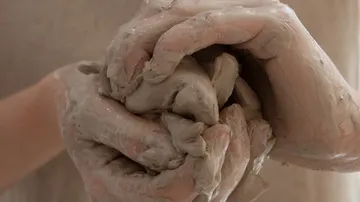 Hands knead moist clay mass in a bright room.