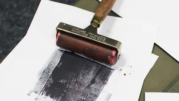 A paint roller glides across printed paper in a studio.