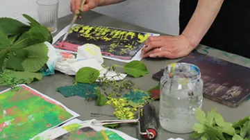 A hand paints on paper surrounded by plants.