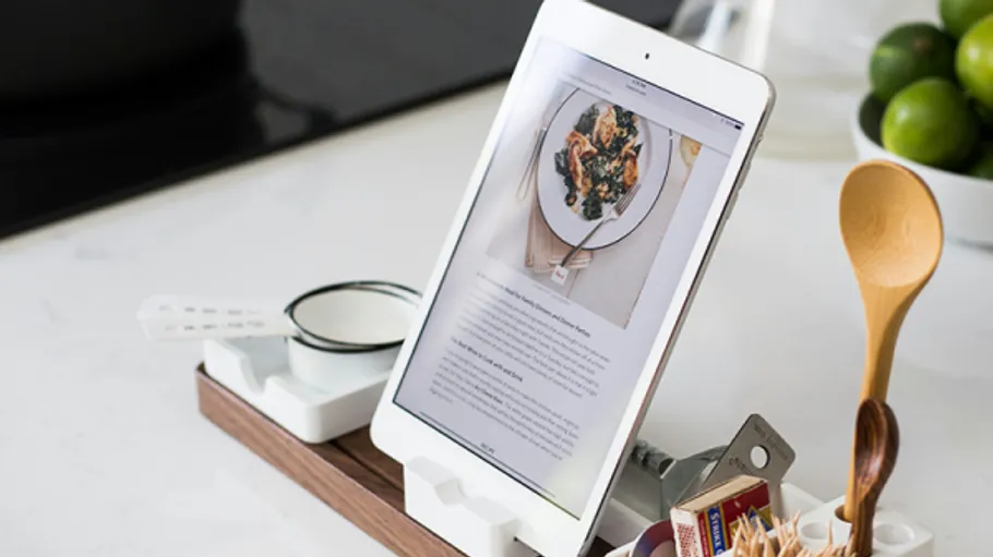 Tablet stands on wooden board, shows recipe, kitchen in background.