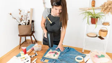 A woman works on jeans with a hair dryer at the table.
