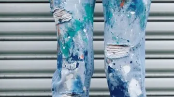 Ripped jeans show splashes of paint against a striped wall.