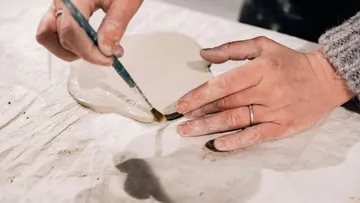 Hände modellieren Ton und zeichnen mit Pinsel auf Oberfläche.