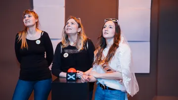 Three women look curiously at a red button.