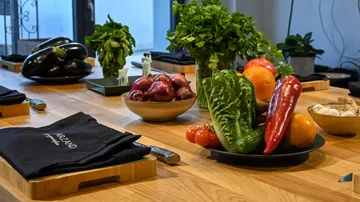 Vegetables and herbs lie on a wooden table in a kitchen.