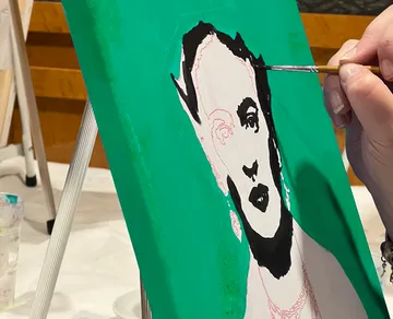 A hand paints a face on a green canvas.