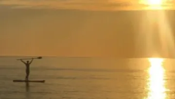 Person stands on a paddleboard, paddles at sunset.