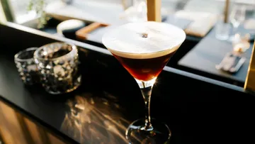 Cocktail glass with dark drink and foam, on table.