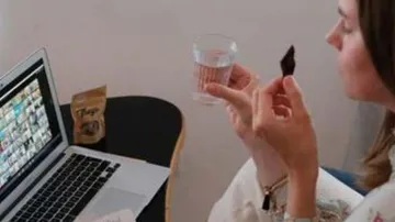 A woman is sitting at the table, drinking water and looking at the laptop.
