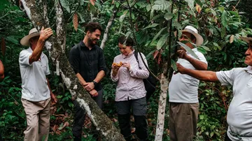 Two people look at a fruit in the jungle with locals.