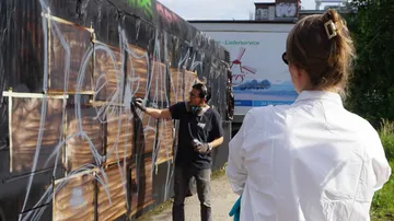An artist sprays graffiti on a wall, watched by someone.