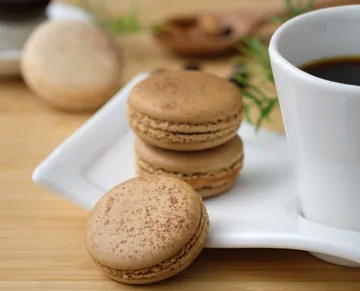 Three macarons lie on a plate next to a cup of coffee.