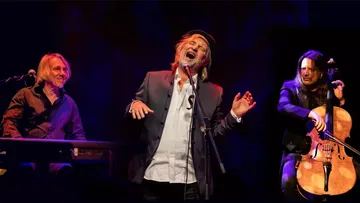 Three musicians perform, sing and play instruments on stage.