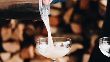 A cocktail is poured into a glass, piles of wood in the background.