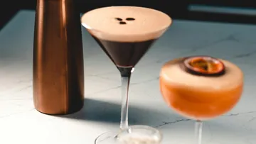 Zwei Cocktails stehen auf einem Tisch neben einem Shaker.