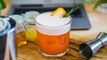 A cocktail is sitting on a table with ingredients and a laptop.